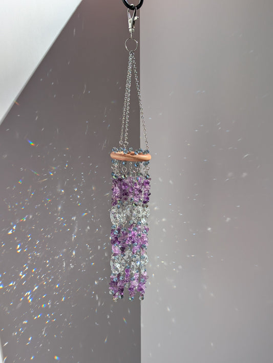Natural amethyst suncatcher with purple gemstone chips and clear crystal prisms hanging from a wooden ring, with rainbow reflections behind it.
